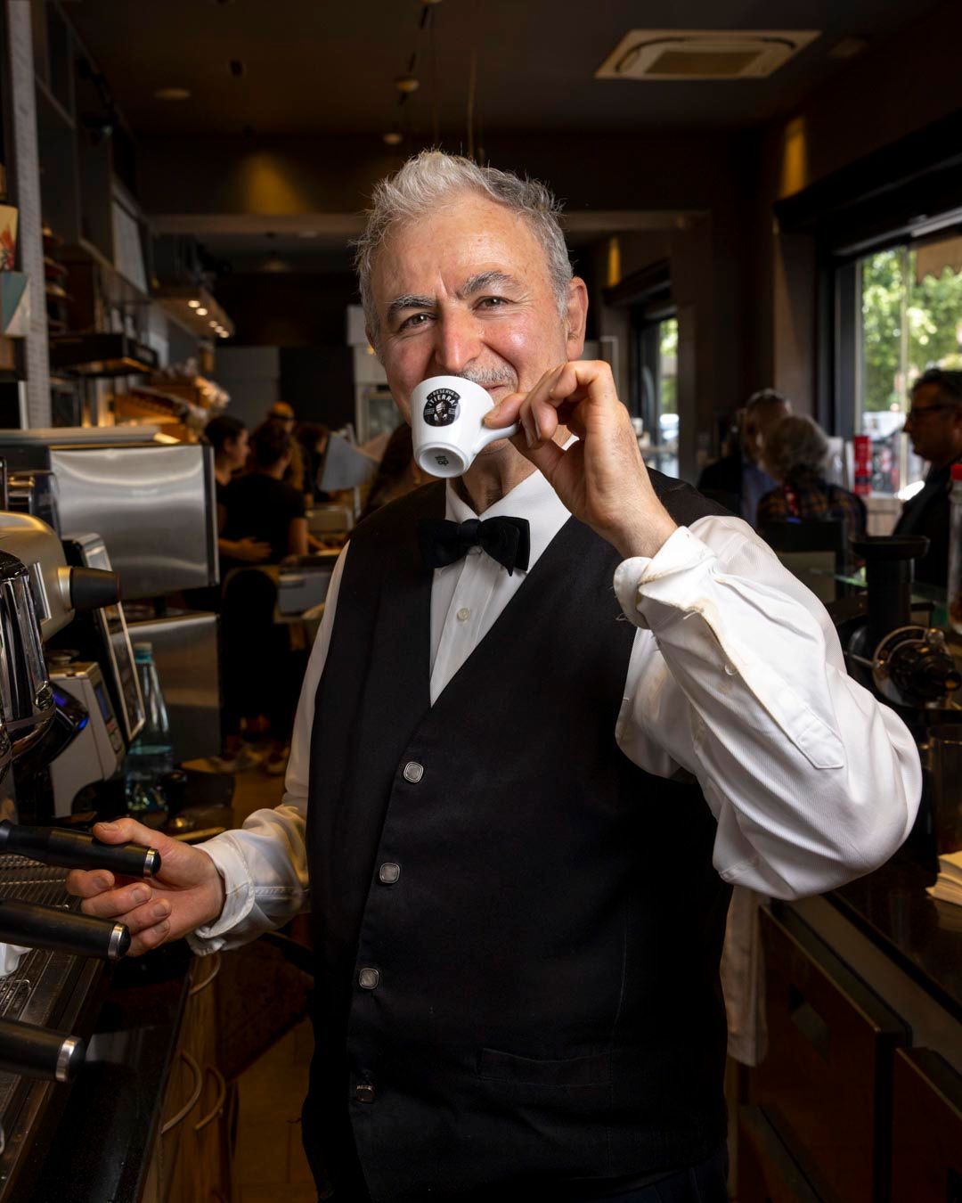 Uomo in gilet e papillon che sorseggia un caffè da una tazza in un bar.