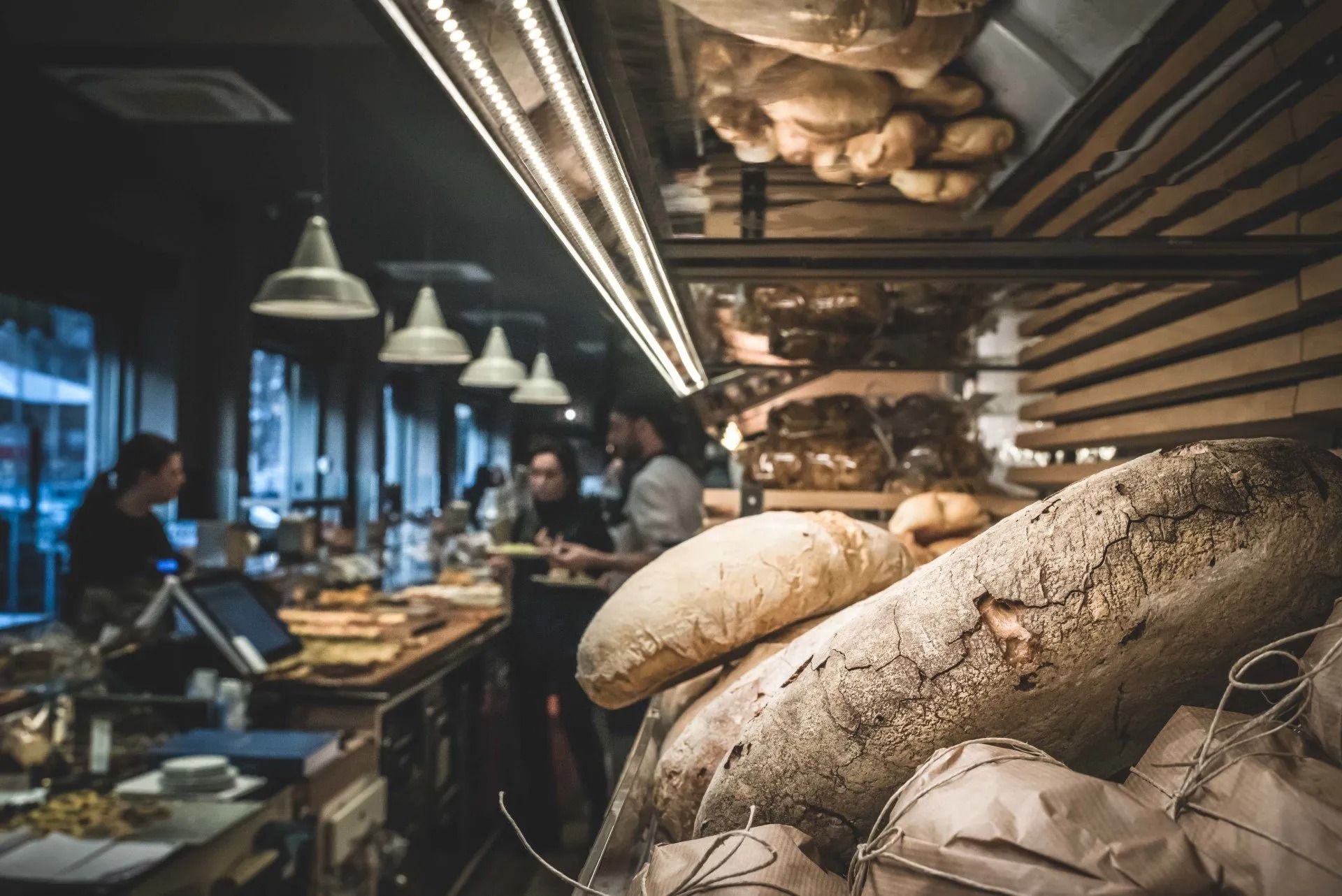 Interno di un panificio con pagnotte di pane fresco in primo piano, clienti e bancone.