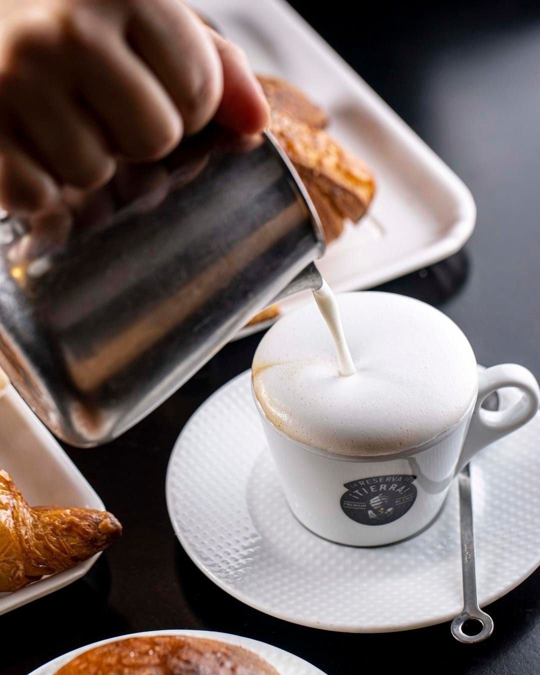 Latte fumante versato in una tazza da cappuccino con un croissant su un piatto.