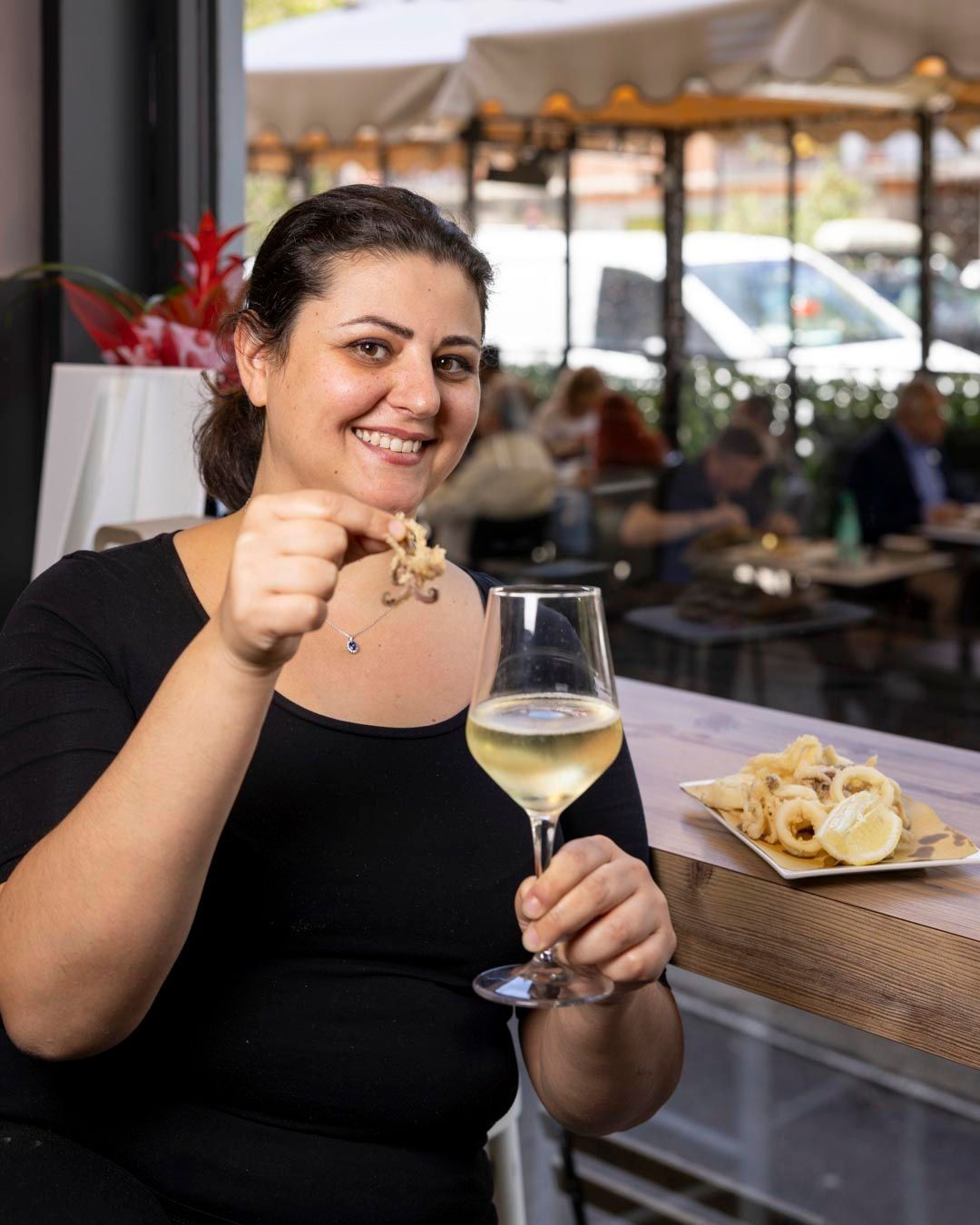 Donna sorridente con in mano calamari fritti e un bicchiere di vino in un ristorante con vista all'aperto.