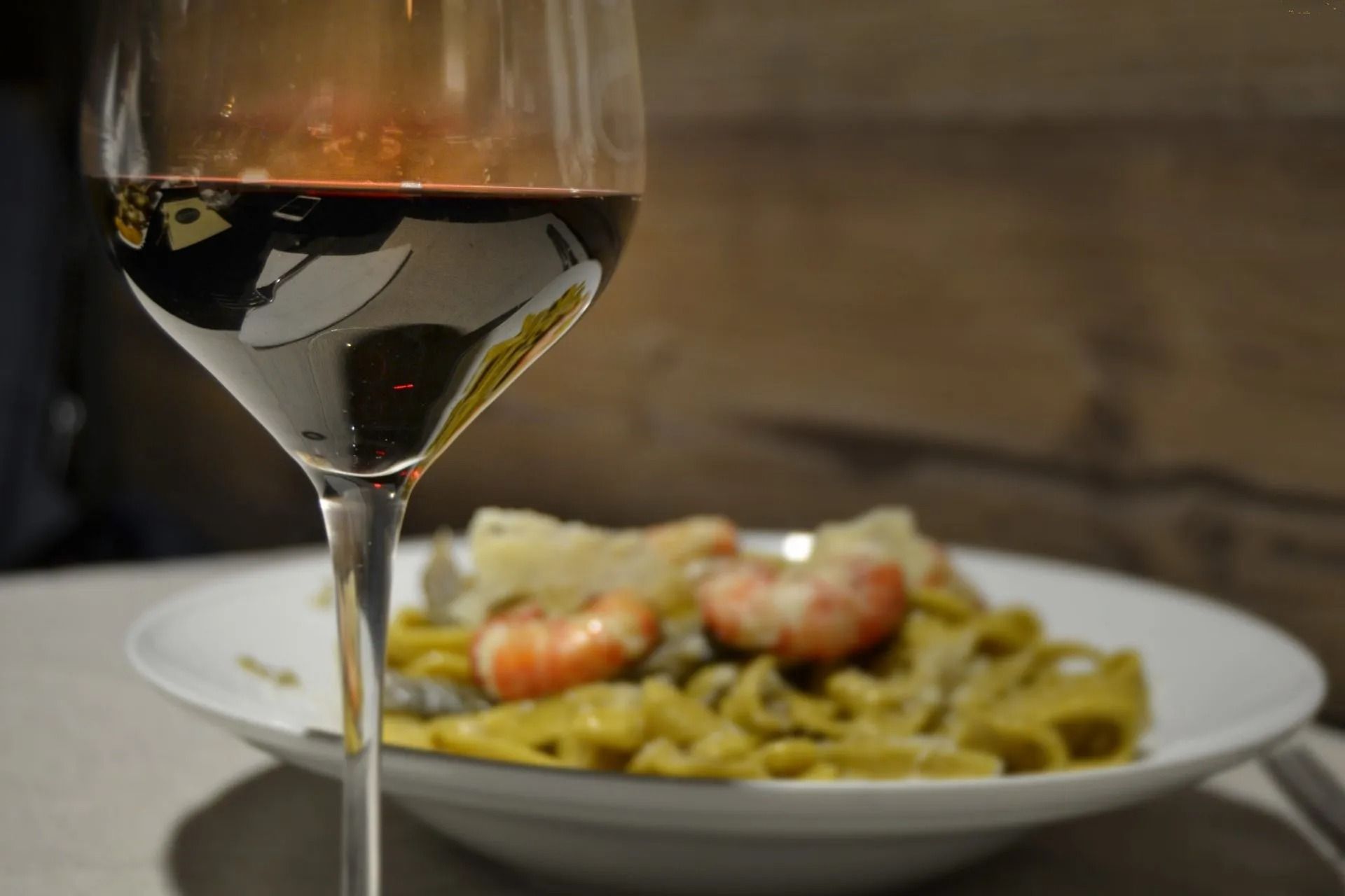 Bicchiere di vino rosso a fuoco, piatto di pasta con gamberi sfocato sullo sfondo.
