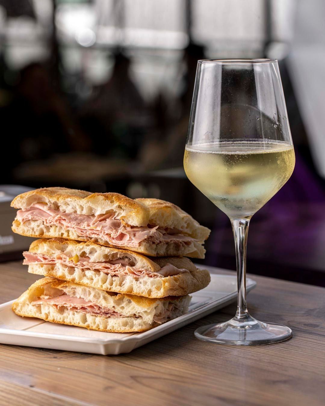 Pila di panini al prosciutto su un piatto bianco con un bicchiere di vino bianco su un tavolo di legno.