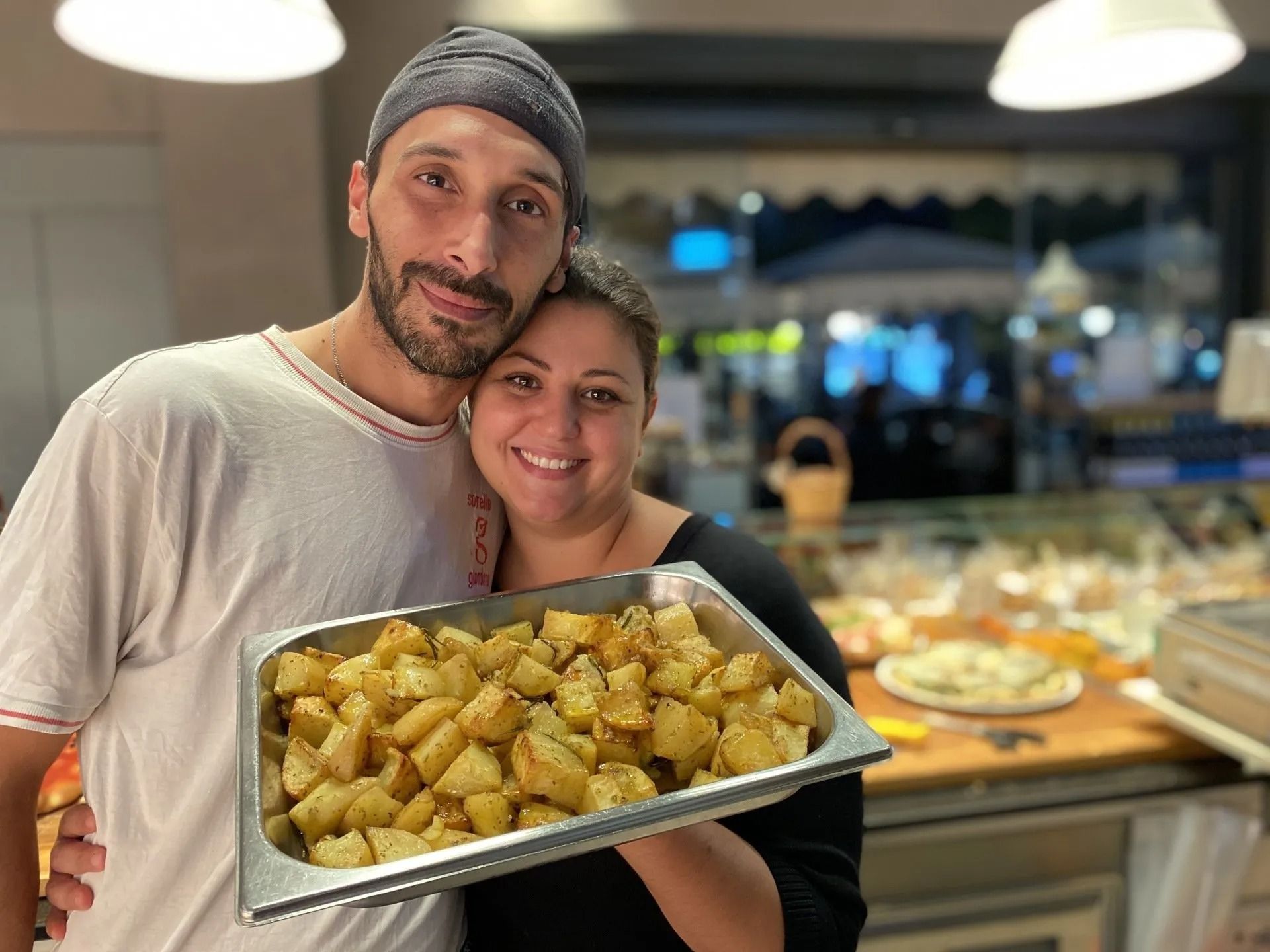 Coppia sorridente con un vassoio di patate arrosto in mano in un negozio di alimentari. Luci calde, vari dolci sullo sfondo.