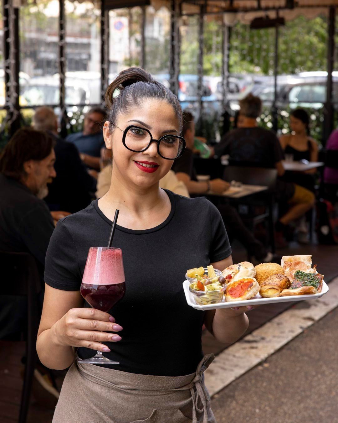 Cameriera che tiene in mano un drink e un piatto di cibo, sorridendo alla telecamera in un bar all'aperto.