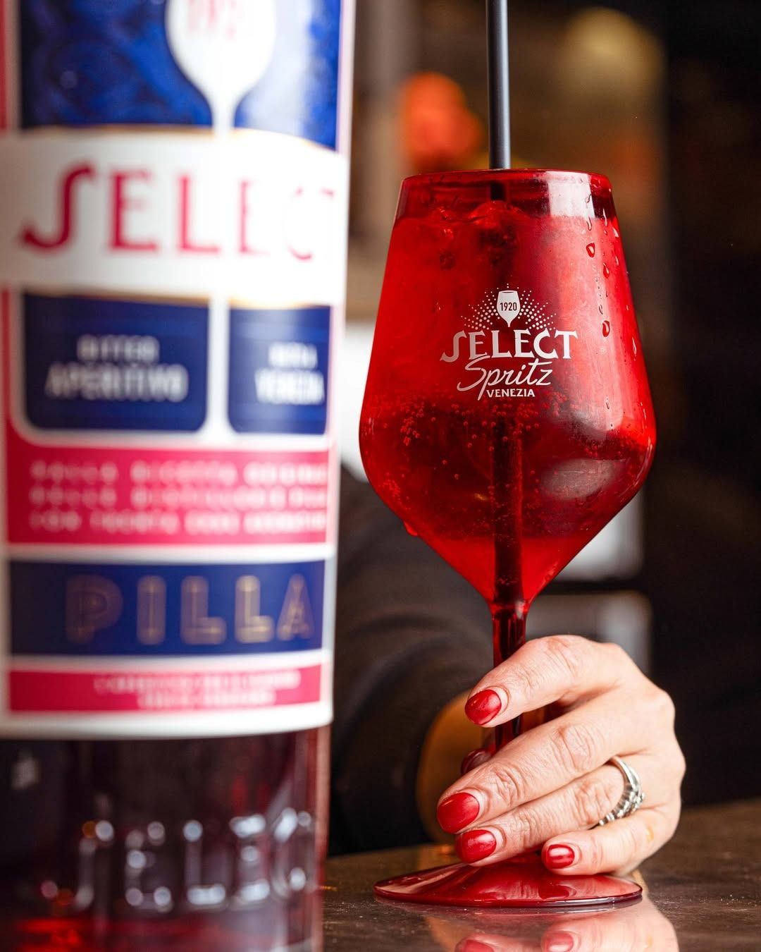 Una mano che regge un bicchiere rosso Select Spritz con accanto una bottiglia di Select Aperitivo.