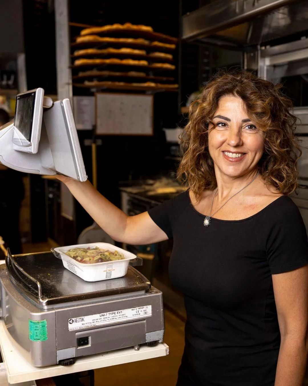 Donna sorridente che pesa il cibo al bancone di una panetteria. Sullo sfondo, pile di pasticcini.
