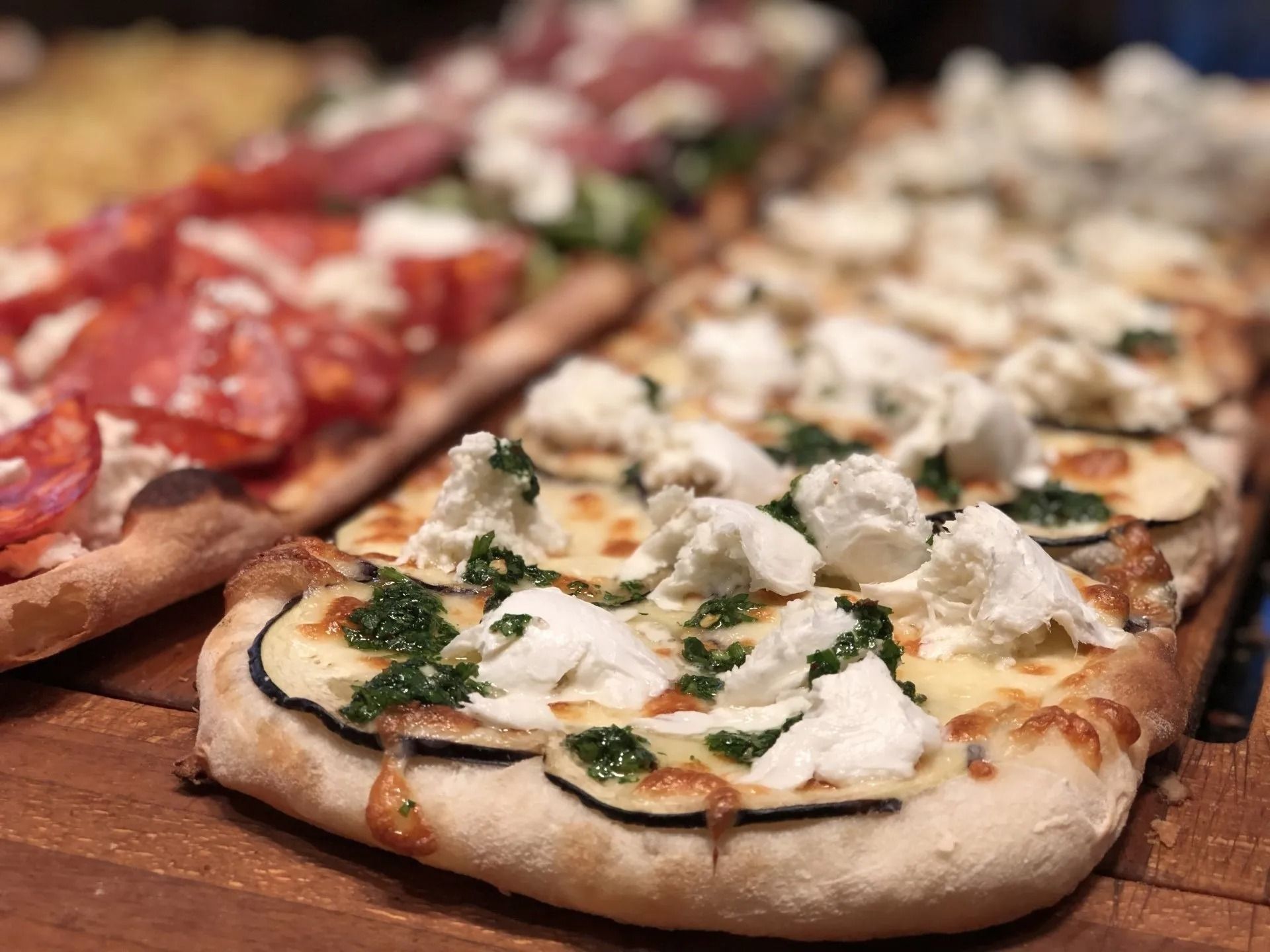 Pizze rettangolari con vari condimenti: pomodoro, chorizo, formaggio di capra, zucchine e pesto.