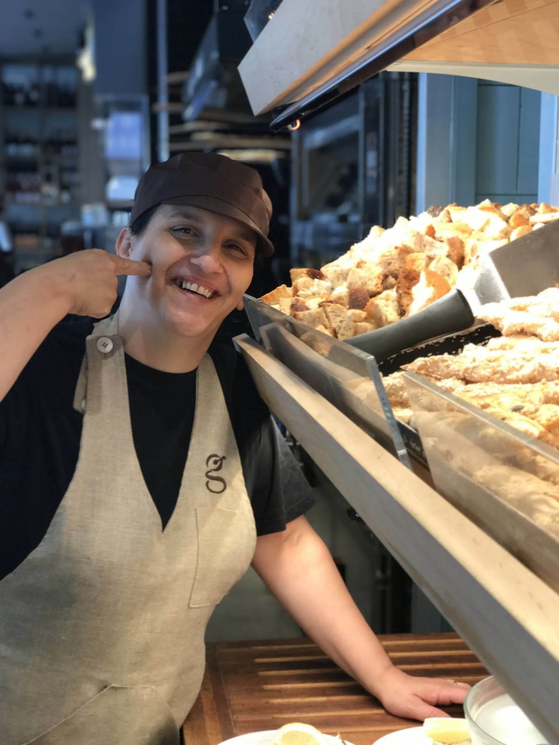 Una persona con grembiule e cappello indica la guancia, sorridendo, vicino all'espositore del pane.