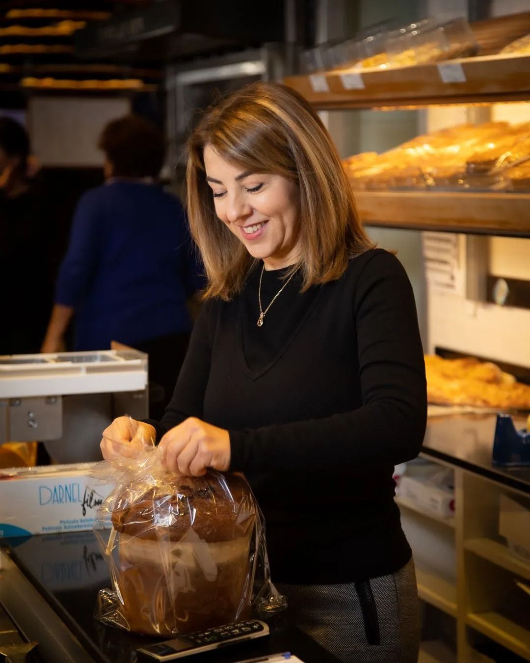 Donna sorridente con maglione nero che confeziona il pane in un panificio, con prodotti da forno esposti sullo sfondo.