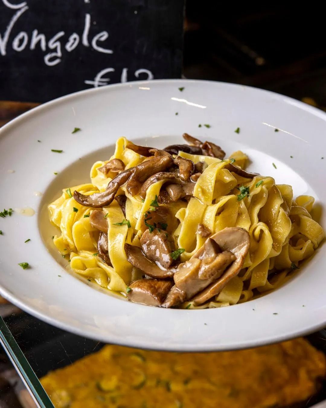 Pasta con funghi in una ciotola bianca, piccoli rametti di prezzemolo, prezzo 12 €.