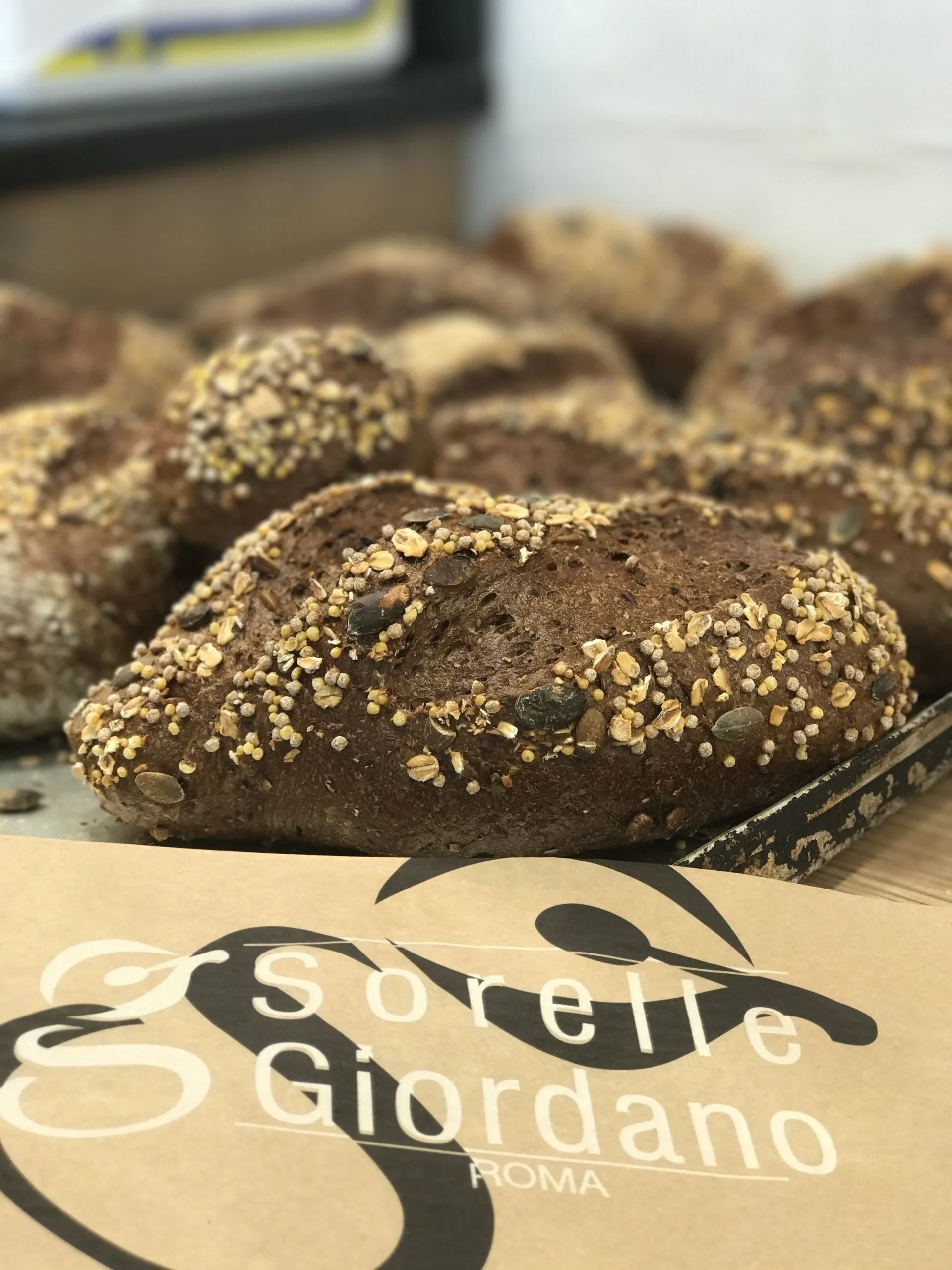 Primo piano di una pagnotta di pane integrale con semi su un vassoio. In primo piano il logo "Sorelle Giordano Roma".