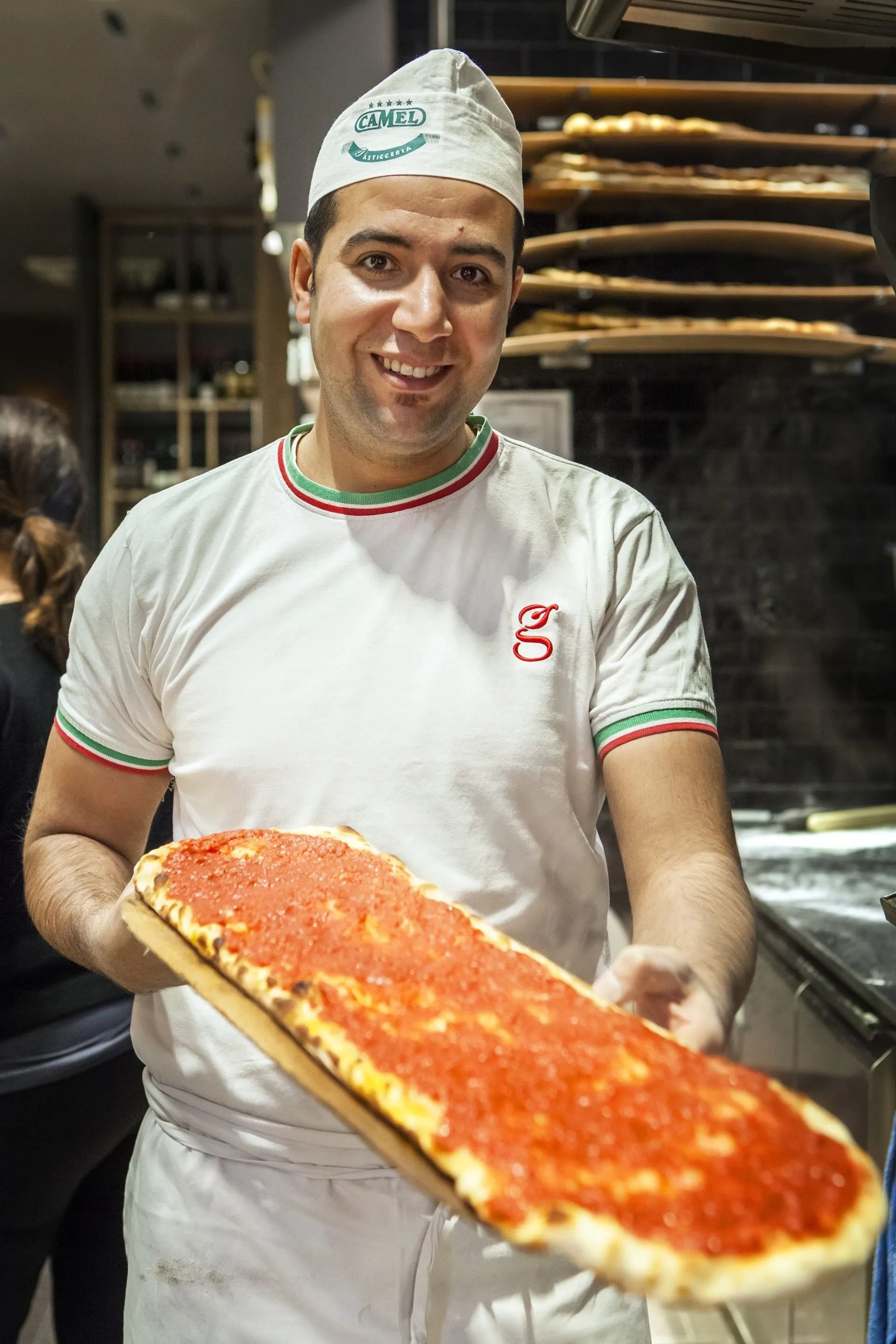 Il pizzaiolo tiene in mano una grande pizza rettangolare con salsa rossa; indossa un cappello bianco e una camicia, ambientazione da ristorante.
