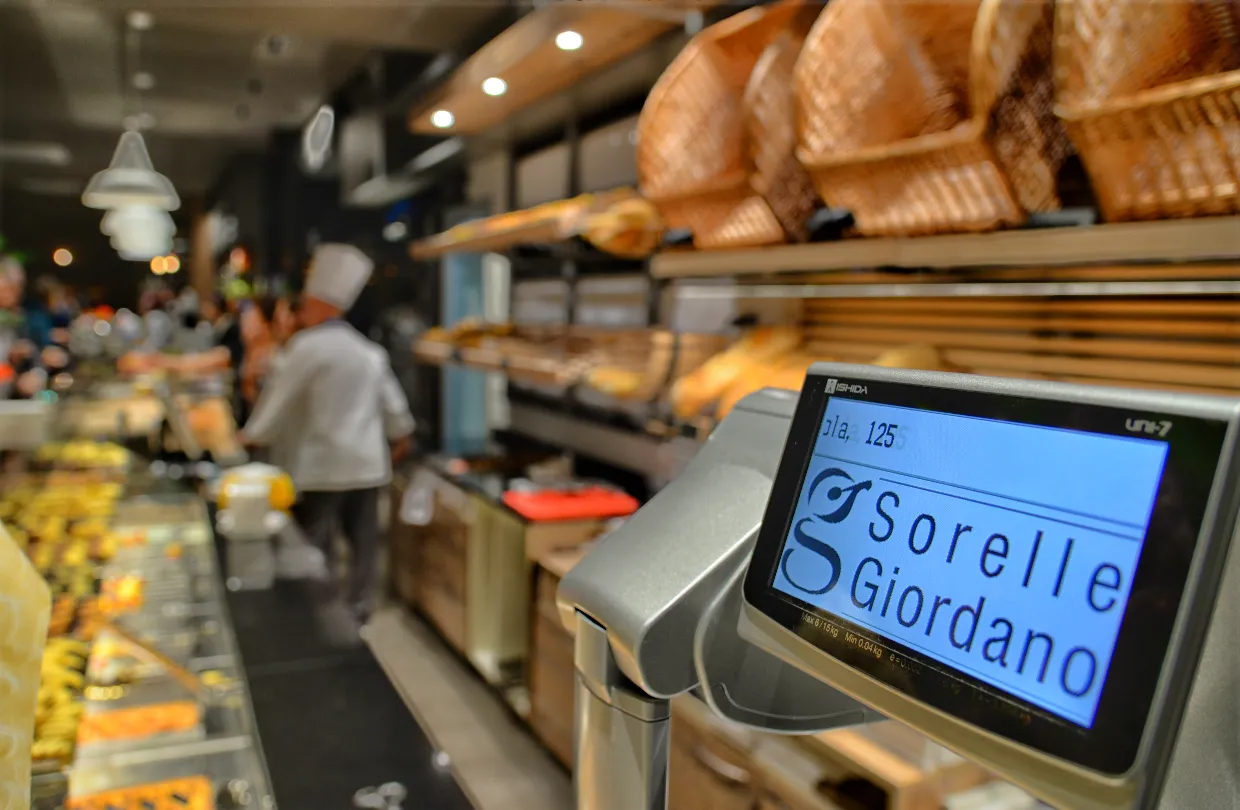 Bancone del panificio con display digitale; sullo sfondo cammina una fornaia in camice bianco; sul display è scritto "Sorelle Giordano".