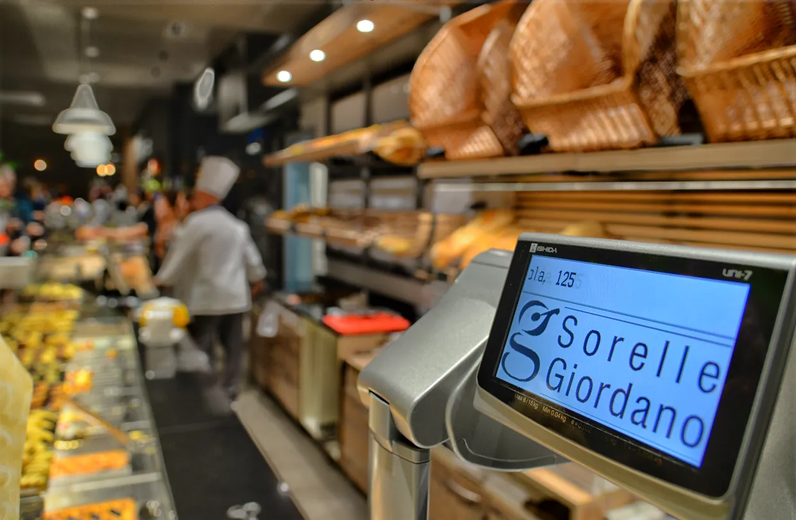 Bancone del panificio con display digitale; sullo sfondo cammina una fornaia in camice bianco; sul display è scritto "Sorelle Giordano".
