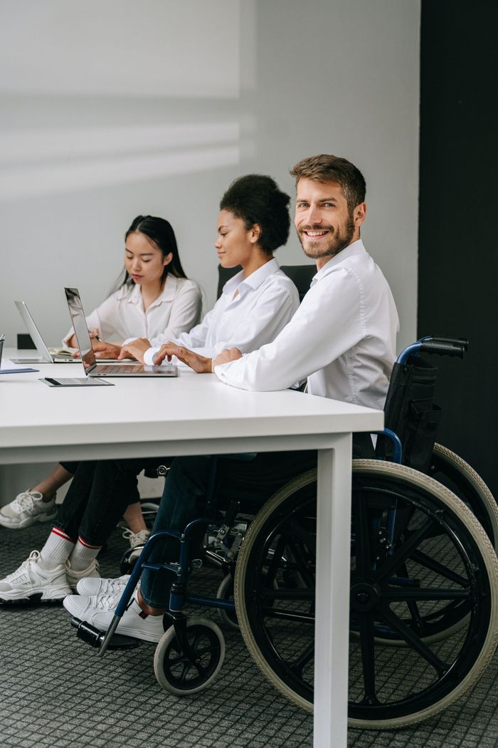 Two women and a man in smart casual wear are in a business meeting. The man who is in a wheelchair turns his head to the side and smiles.