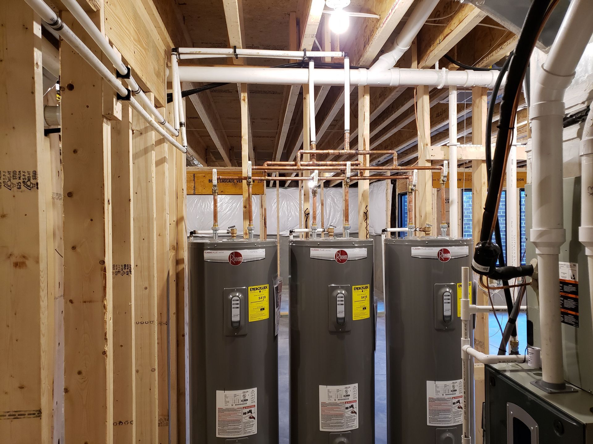 Three tall, gray water heaters in a wood-framed room, with exposed pipes and wiring overhead.