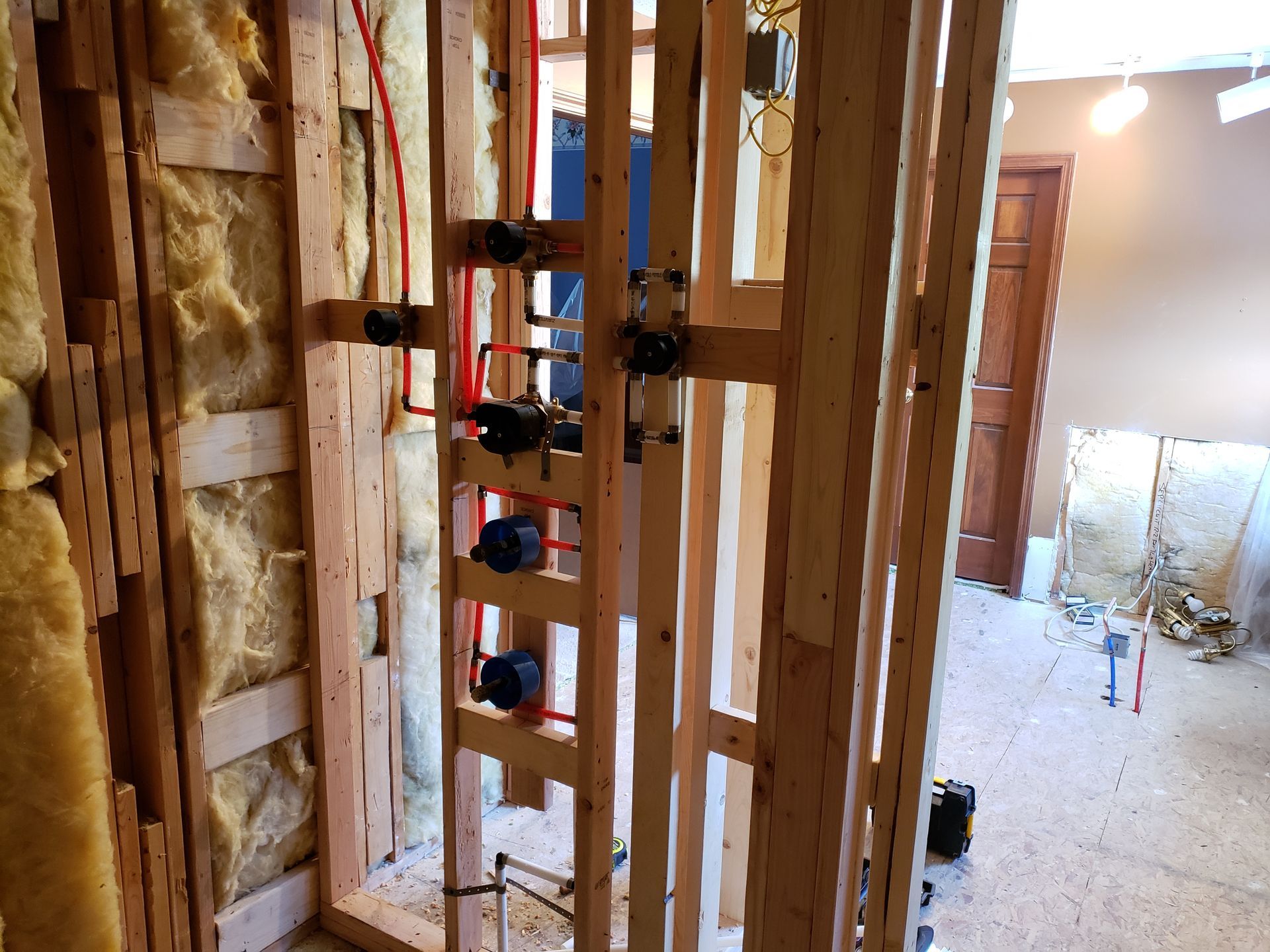 Framed wooden plumbing in a wall, with red PEX pipes, black valves, and insulation.