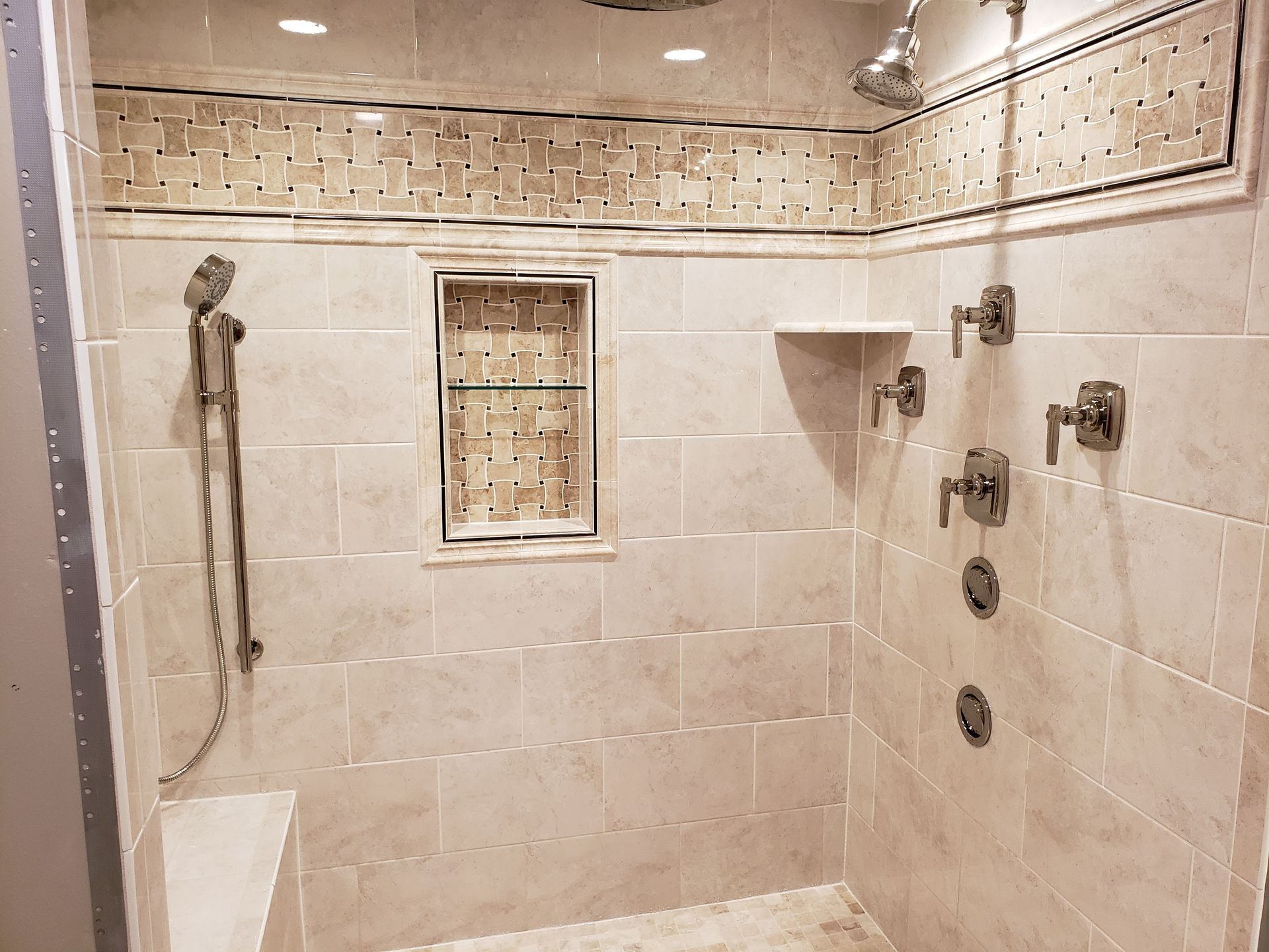 Tiled shower with handheld and overhead showerheads, built-in shelf and niche, beige and brown tones.