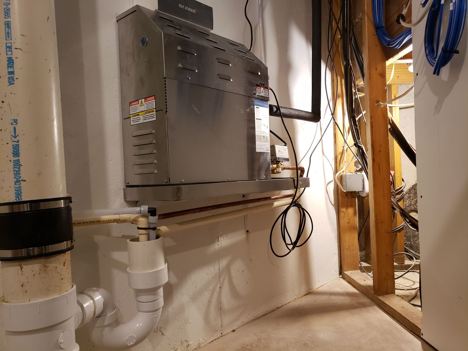 Silver water heater mounted on a white wall near plumbing pipes and exposed wires.