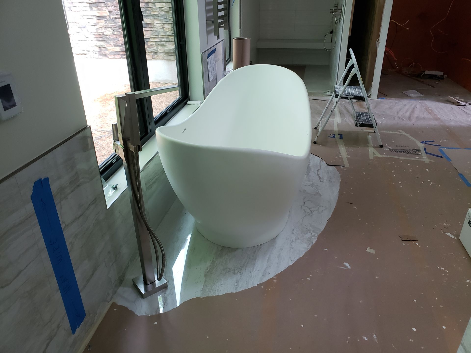 White, oval bathtub in a room under construction, near a window and with a chrome faucet.