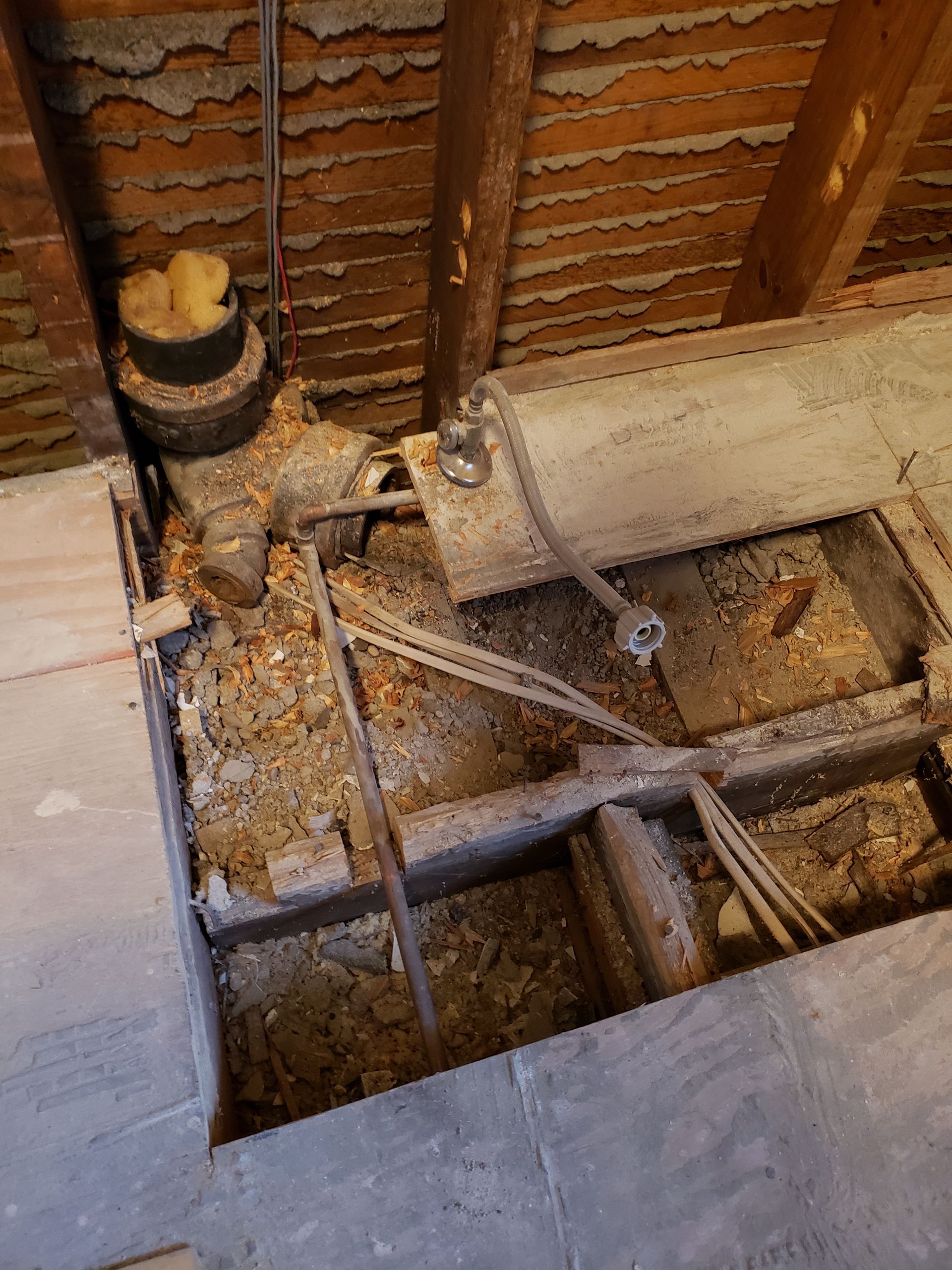 Open floorboards reveal pipes, wires, and debris in an unfinished interior space, likely an attic or basement.