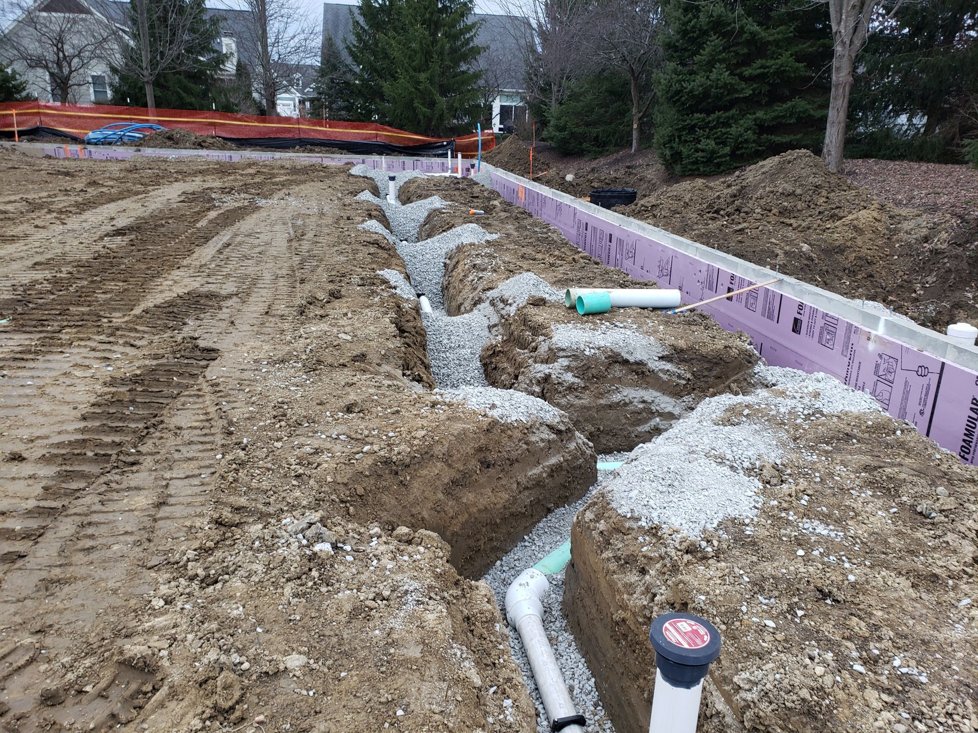 Trenches with piping and gravel for drainage, next to a building's foundation with pink insulation.