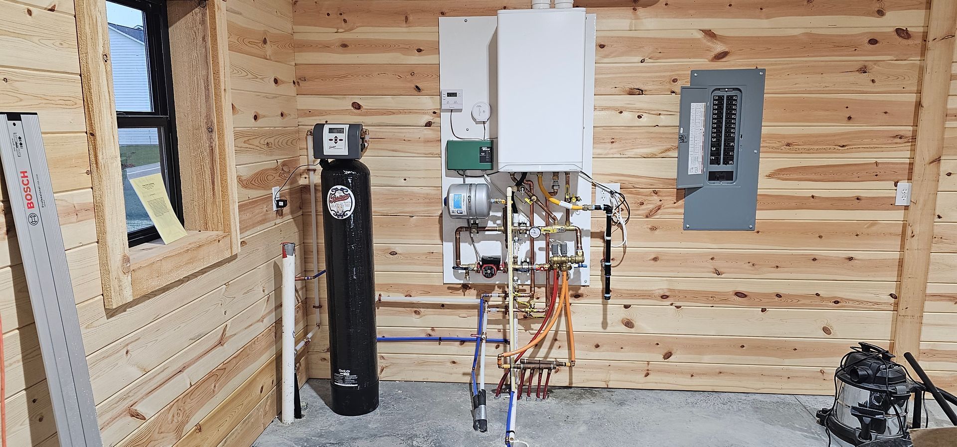 Interior of a wooden cabin with a water softener, plumbing, and electrical panel visible on the wall.