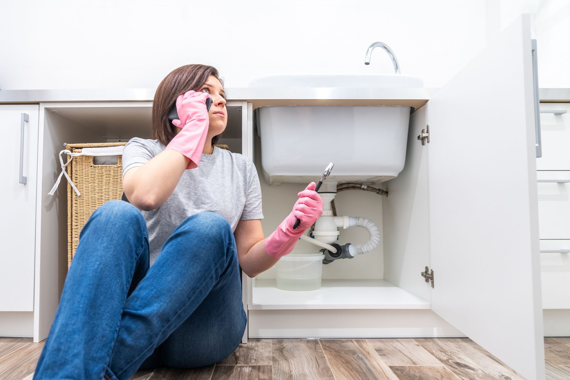 A woman on the phone by a leaking sink is calling for emergency plumbing services.