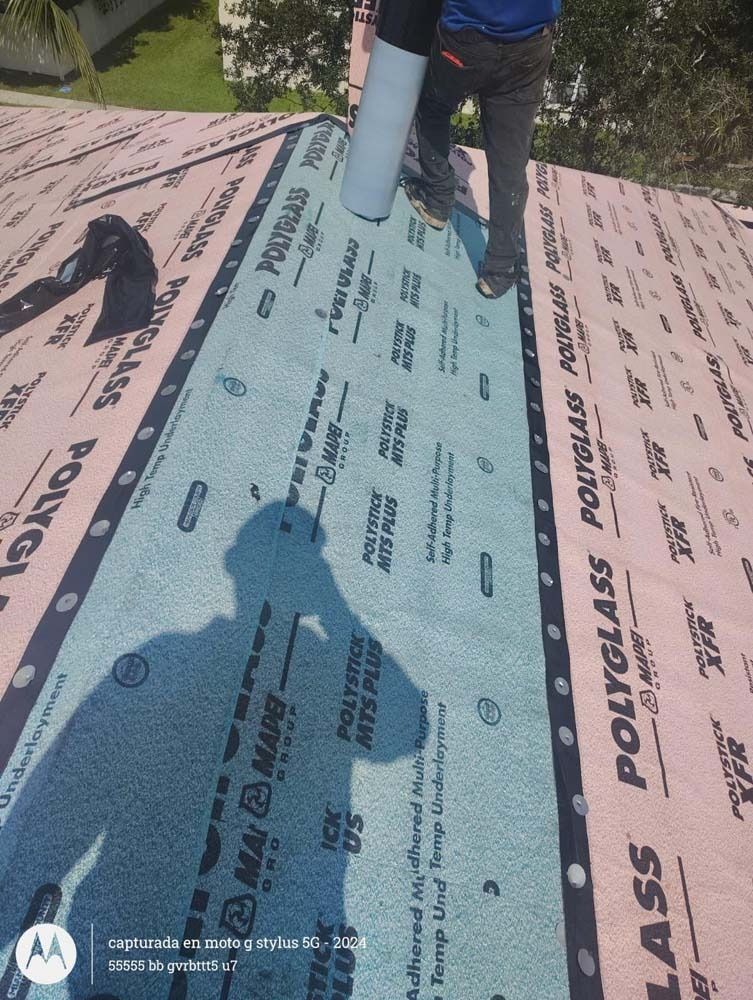 A person in work pants installs rolls of Polyglass roofing underlayment on a residential sloped roof.