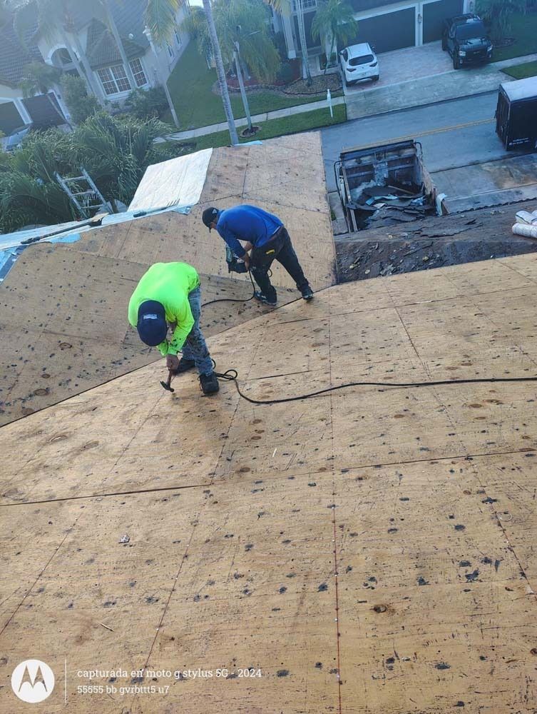 Two construction workers in high-visibility gear install plywood sheathing on a sloped residential roof.