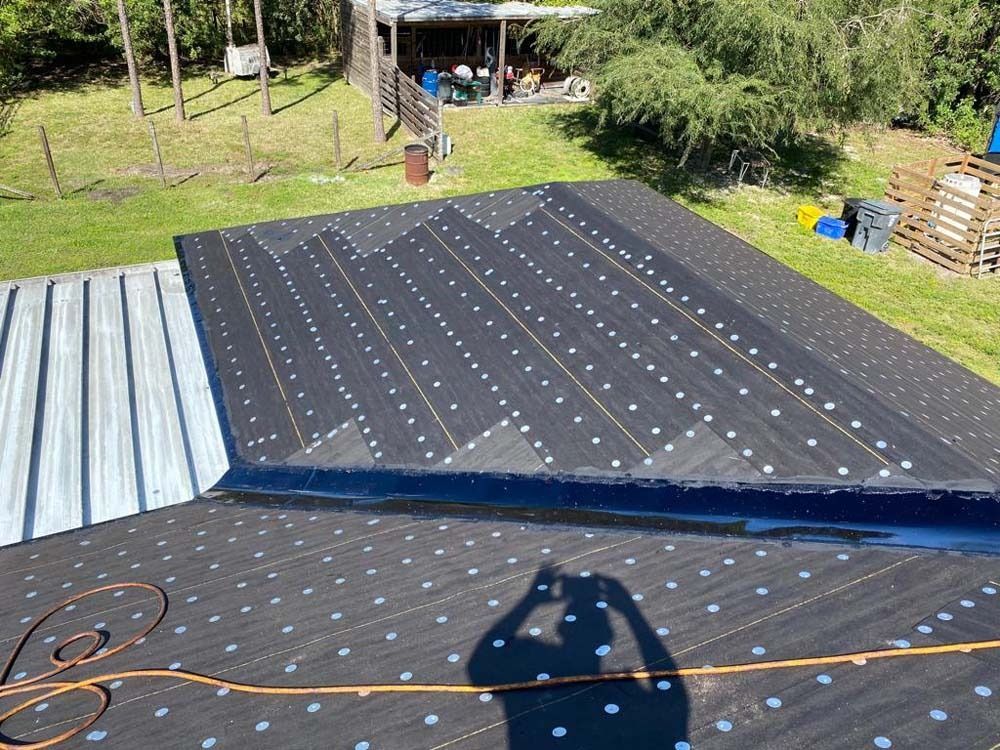 An overhead view of a roof under construction covered in black underlayment, transitioning to existing metal roofing.