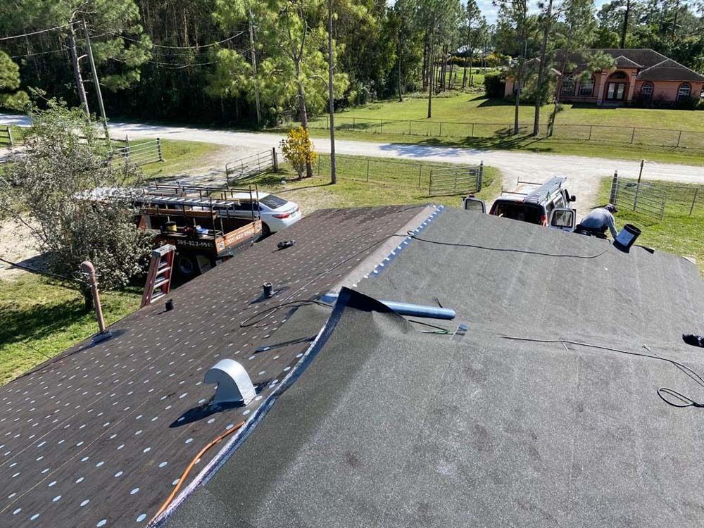 Workers repairing a residential roof on a sunny day, with a vehicle and equipment visible in the yard below.