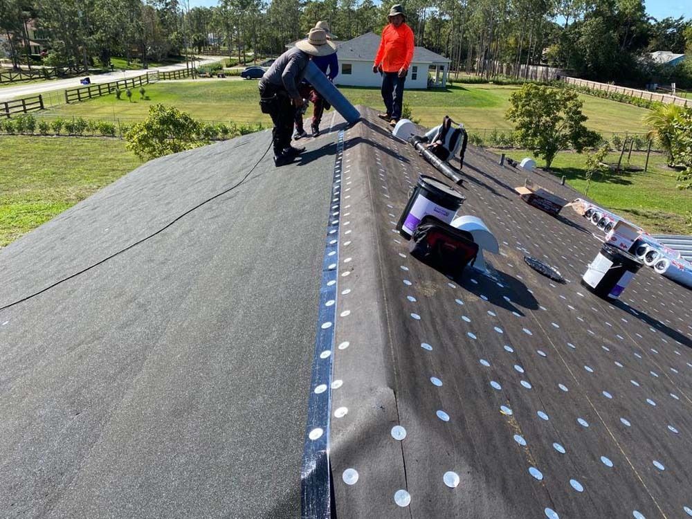 Two workers in hard hats install black roofing underlayment on a steep, sunny residential roof.