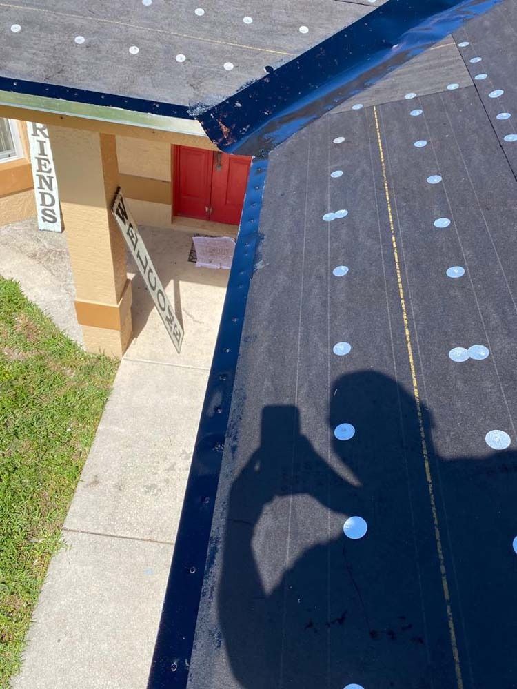 A first-person view from a roof under construction, showing black underlayment with metal cap nails and a red front door below.