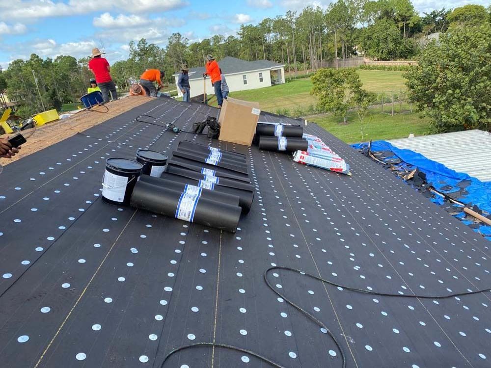 A crew installs black synthetic roofing underlayment on a house roof with building materials scattered nearby.