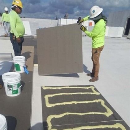 Workers in high-visibility gear install insulation boards on a roof, applying adhesive in a serpentine pattern.
