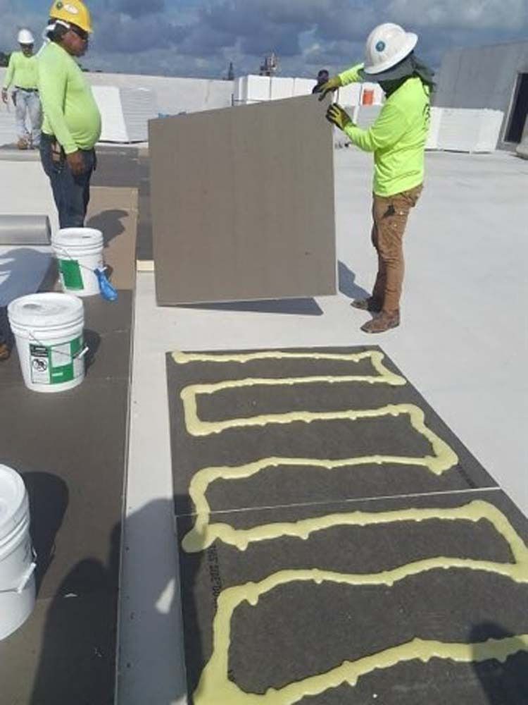 Construction workers in safety gear applying adhesive and installing insulation panels on a flat roof.
