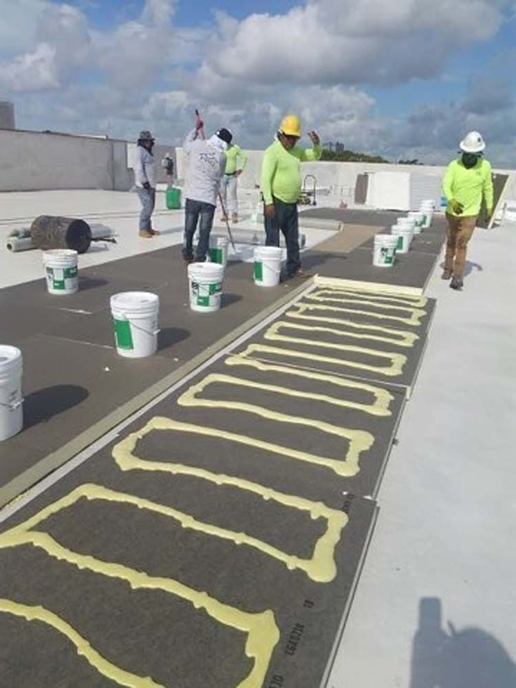 Roofers in neon safety gear applying adhesive patterns to black insulation boards on a flat roof.