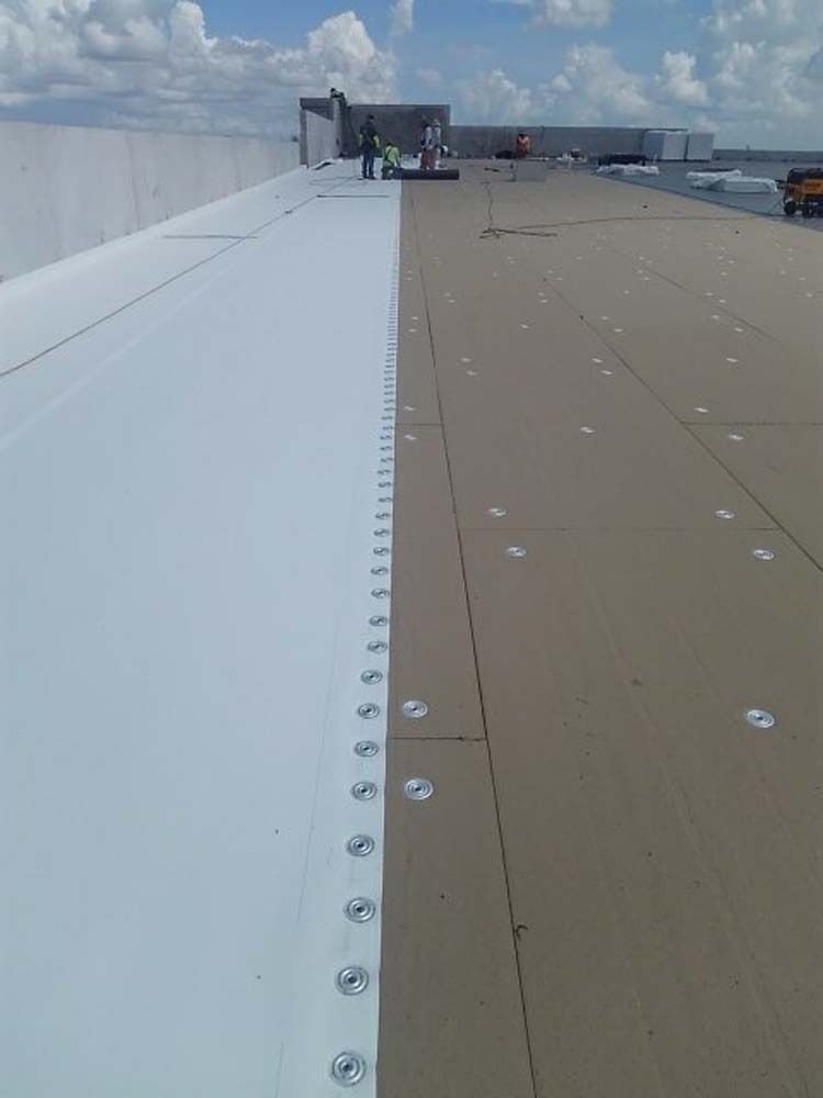 A construction site showing a white membrane being mechanically fastened to a roof surface with a line of metal plates.