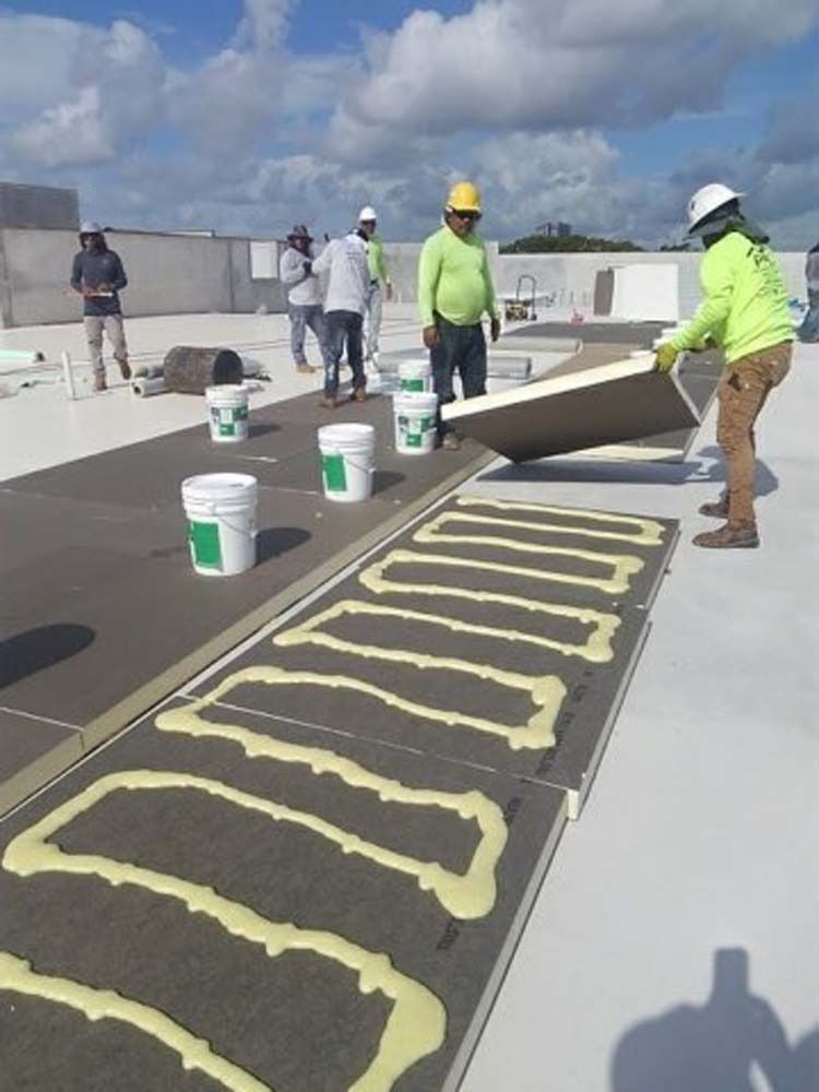 Workers in high-visibility gear install roofing insulation boards onto a white rooftop using adhesive foam beads.