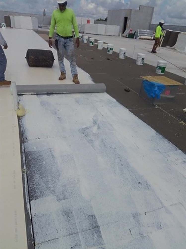 Construction workers in high-visibility gear install white roofing membrane over a dark base on a flat building rooftop.