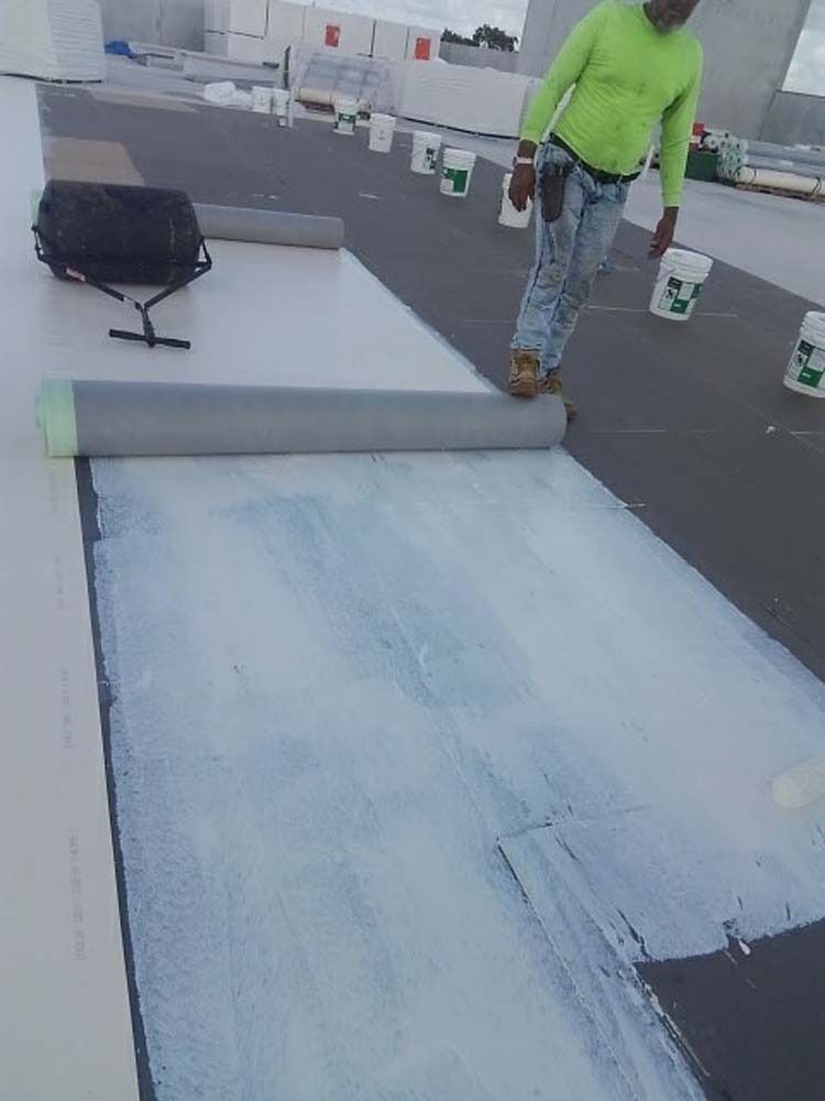 A worker in a neon green shirt rolls out a gray roofing membrane over a white adhesive-coated surface on a flat rooftop.