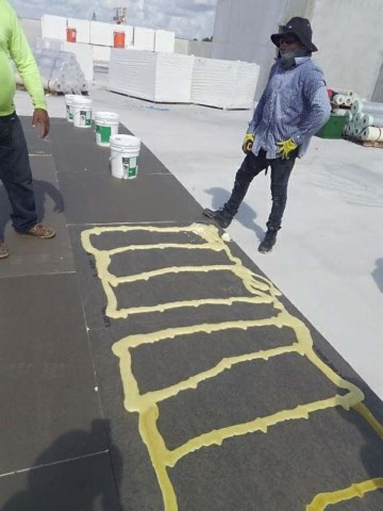 Two workers on a flat roof next to buckets and adhesive beads applied in a grid pattern to install roofing material.