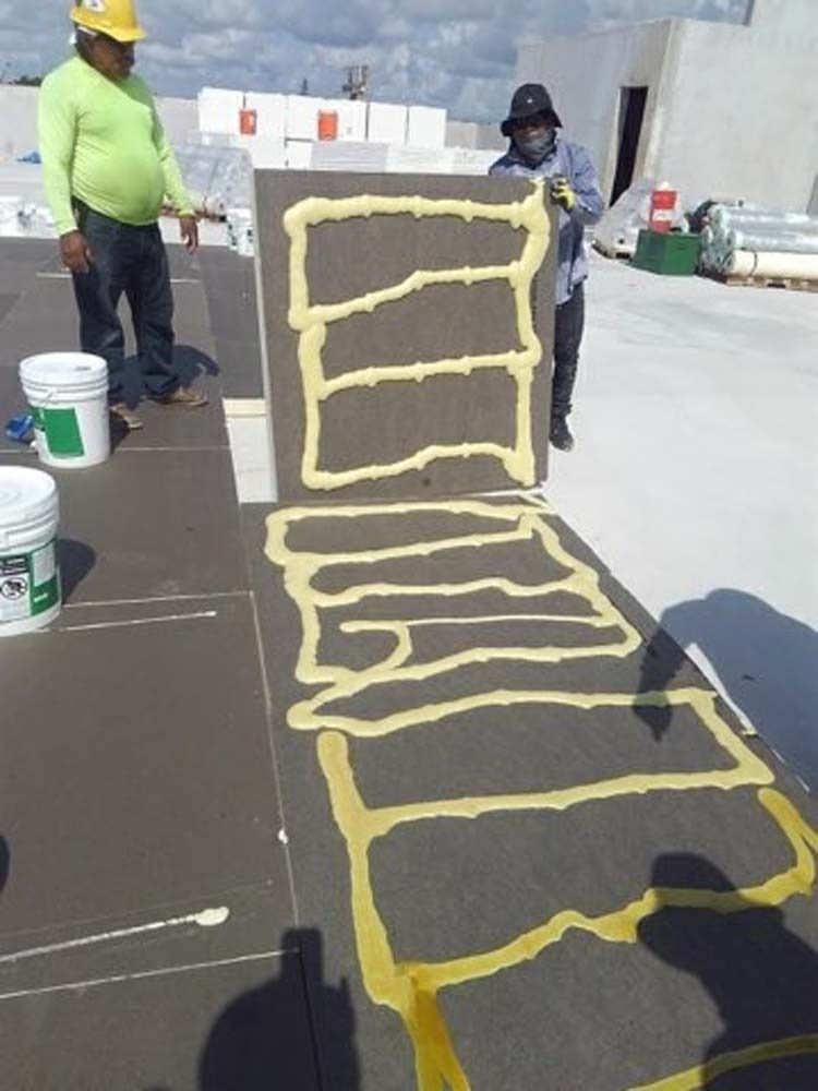 Two workers on a flat roof applying adhesive foam to a board for insulation installation.