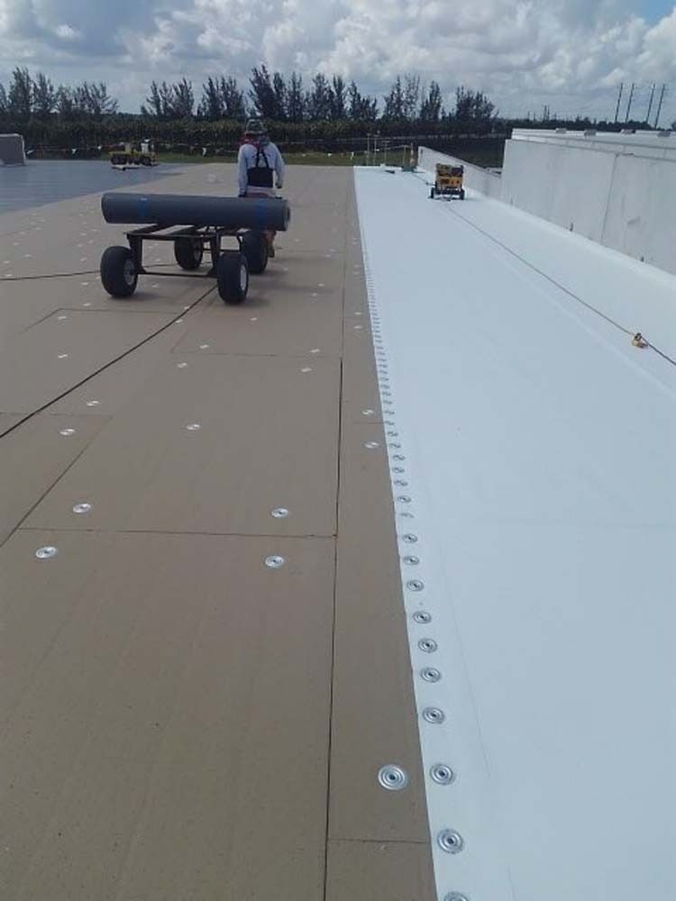 A worker operates a rolling tool on a flat roof being installed with brown insulation boards and white membrane sheets.