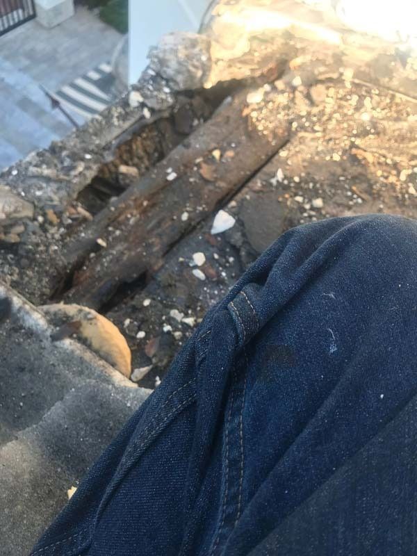 A close-up view of a person’s blue-jeaned lap next to a section of a crumbling stone wall or roof edge with exposed wood.
