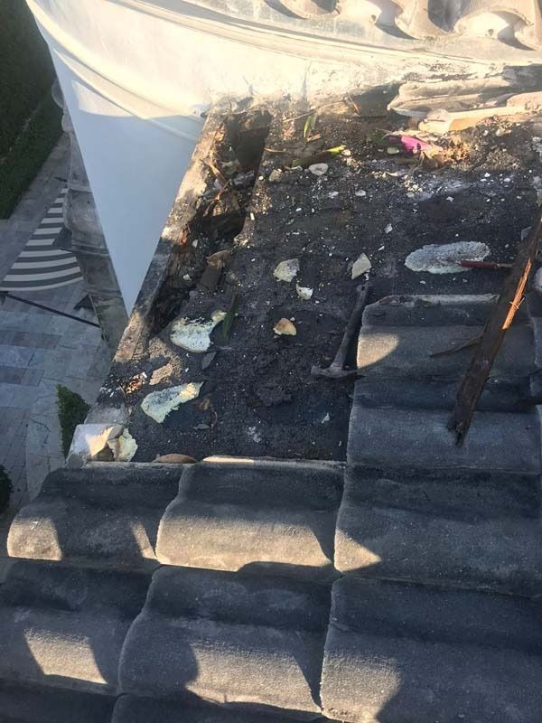 Damaged roof section with missing tiles, exposed dark underlayment, debris, and a hammer, viewed from above.