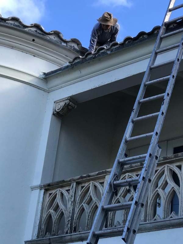 A person in a hat works on the edge of a roof, next to a metal ladder leaning against a building with stone balconies.