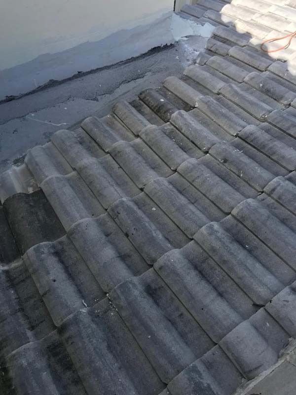 A grey tiled roof with a damaged flashing area along the wall, showing signs of wear and gaps in the sealant.