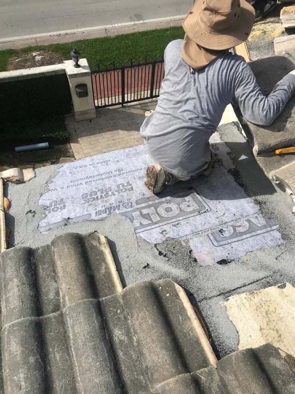 A worker wearing a sun hat kneeling on a residential roof, repairing the underlayment beneath removed concrete tiles.